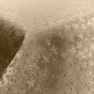 Puede incluir: Un mantel beige con un sutil patrón de estrellas plateadas. La tela tiene una textura ligeramente texturizada, casi brillante.
