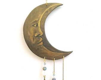 Pode incluir: Uma lua crescente dourada com um rosto pintado nela pendura em uma corrente dourada. Três estrelas douradas pendem da parte inferior da luna, cada uma com uma conta branca e uma conta azul iridescente em uma corrente dourada.