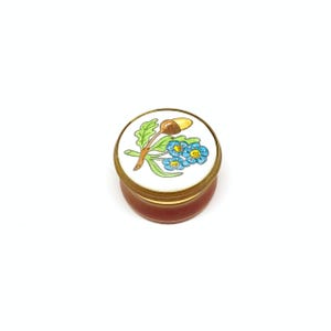 Puede incluir: Una pequeña caja redonda de esmalte blanco con tapa dorada. La tapa presenta un diseño pintado a mano de una flor azul con una bellota marrón en un tallo verde.