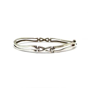 May include: A silver-toned bracelet with an intricate, linked design. The bracelet features a series of interconnected, curved bars and decorative infinity-shaped links. The clasp is visible on the right side. The jewelry is set against a white background.