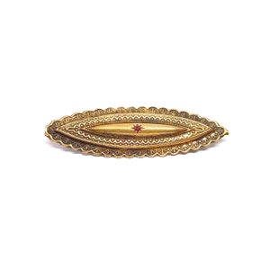 Könnte beinhalten: Eine goldfarbene Brosche in ovaler Form mit filigranen Details. In der Mitte der Brosche befindet sich ein kleiner roter Edelstein.