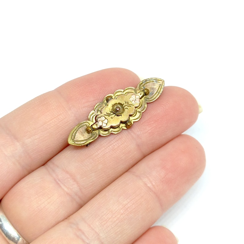 May include: A gold-toned brooch with a floral design and a small, round, red stone in the center. The brooch is in the shape of a long oval.