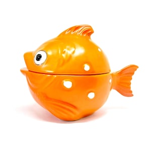 May include: Orange ceramic fish-shaped container with a lid. The container has a round body, a tail fin, and a dorsal fin. It features white circular cutouts and a black and white eye.