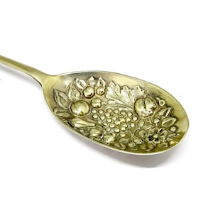 Könnte beinhalten: Ein antiker, silberfarbener Löffel mit einem detaillierten geprägten Design aus Früchten und Blättern. Die Löffelschale zeigt Trauben, Äpfel und florale Elemente, die ein dekoratives und verziertes Aussehen erzeugen. Der Löffel ist vor einem weißen Hintergrund platziert.
