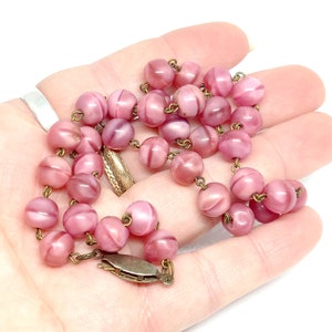 May include: A pink glass bead necklace with a brass clasp. The beads are round and have a swirl pattern.