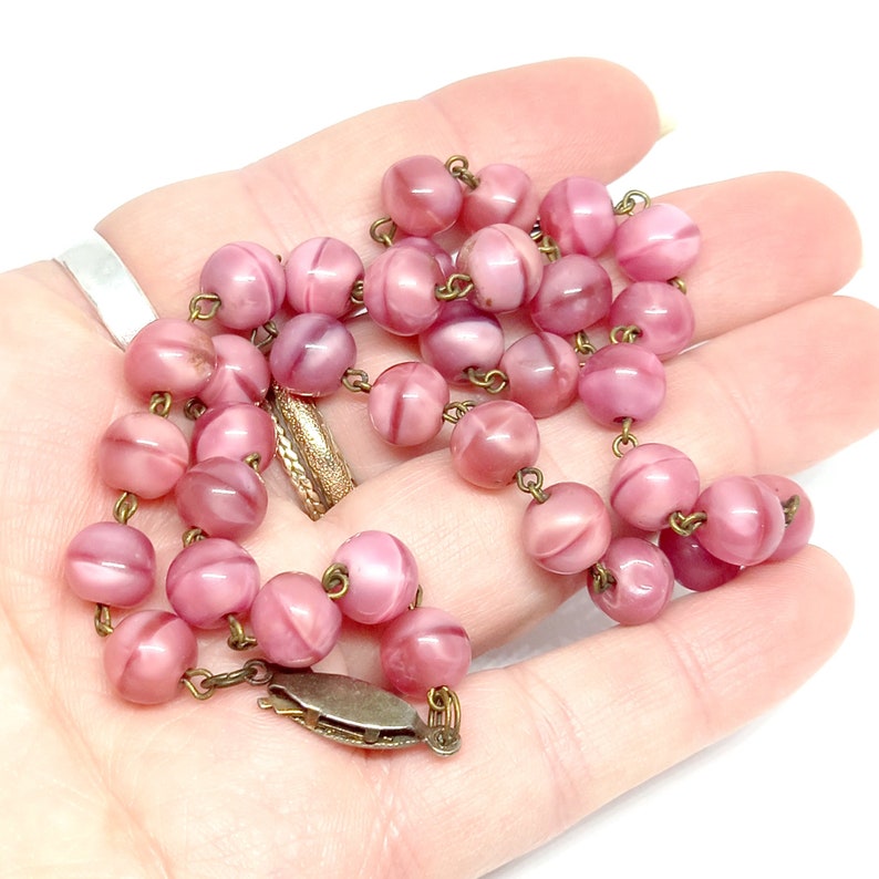 May include: A pink glass bead necklace with a brass clasp. The beads are round and have a swirl pattern.