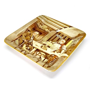 May include: Square ceramic ashtray with a vintage illustration depicting a pub scene. The image features figures at a bar, with the word "BASS" visible. The ashtray has a gold trim and a light brown color scheme.
