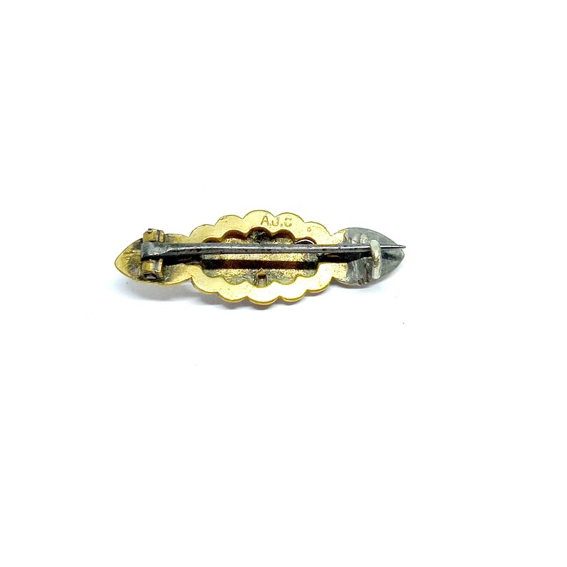 May include: A gold-toned brooch with a scalloped edge and a silver-toned pin. The brooch has the inscription "A.J.C." on the back.