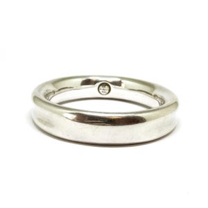 May include: A silver ring with a smooth, curved design. The ring has a polished finish and a small, circular marking on the top. The ring is set against a white background.