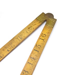 Puede incluir: Una regla plegable de madera vintage con bisagras de latón. La regla está marcada con números negros y líneas de medición. La madera tiene un tono cálido y envejecido, y la regla está parcialmente abierta.