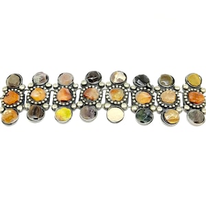 Può includere: Un braccialetto d'argento con una fila di cabochon rotondi di pietre preziose multicolori. Le pietre sono incastonate in castoni d'argento e sono circondate da piccole perle d'argento.