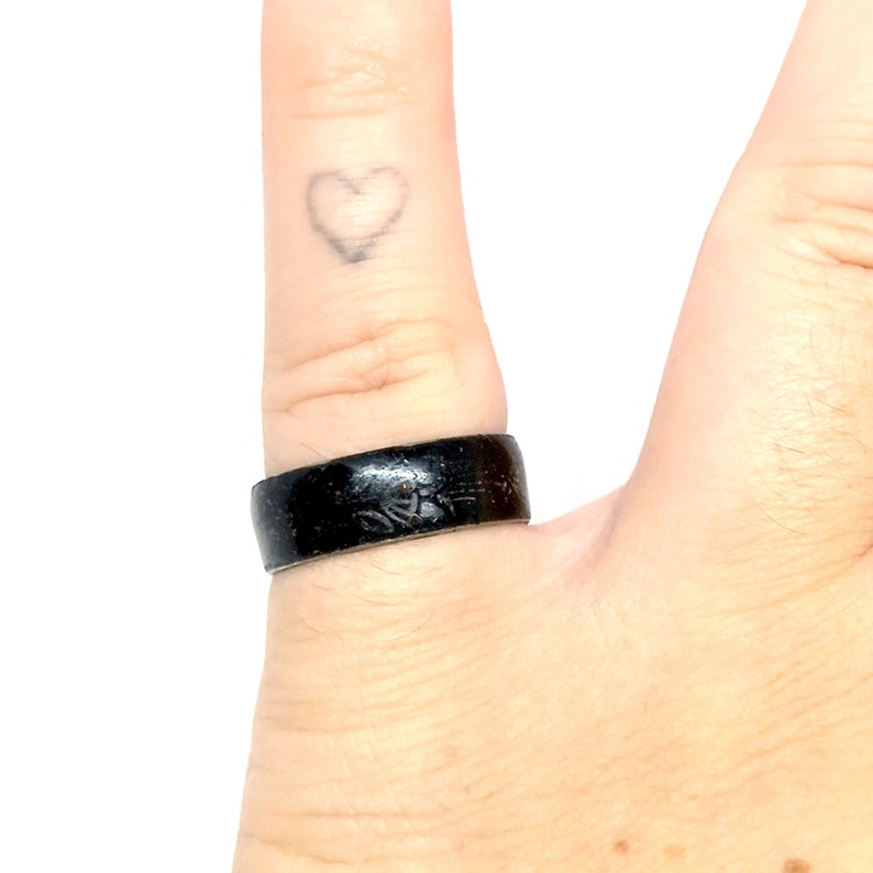 K&ouml;nnte beinhalten: Ein schwarzer Ring mit Holzmaserung. Der Ring wird am Finger einer Person getragen.