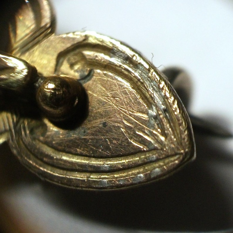 May include: Close-up of the back of a gold-toned heart-shaped pendant with a smooth, slightly textured surface. The pendant has a small, round loop at the top for attaching a chain.