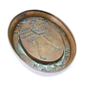 May include: Oval-shaped bronze ashtray with a raised central design. The design features a walking gentleman with a top hat and cane, and the text "BORN 1820 STILL GOING STRONG". The ashtray has a raised rim.