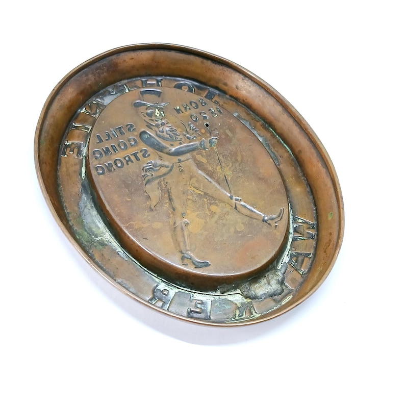 May include: Oval-shaped bronze ashtray with a raised central design. The design features a walking gentleman with a top hat and cane, and the text "BORN 1820 STILL GOING STRONG". The ashtray has a raised rim.