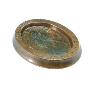 May include: Oval-shaped bronze ashtray with a raised rim. The ashtray has an aged patina and features the text "STILL GOING STRONG" and "WWI" engraved on the surface. The ashtray is a vintage item.