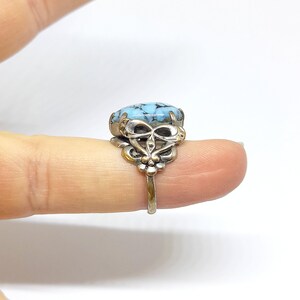 Puede incluir: Un anillo plateado con una piedra ovalada de color azul claro con vetas azul oscuro. El anillo tiene un diseño de filigrana ornamentado alrededor de la piedra y en la banda. El anillo se muestra en un dedo sobre un fondo blanco.