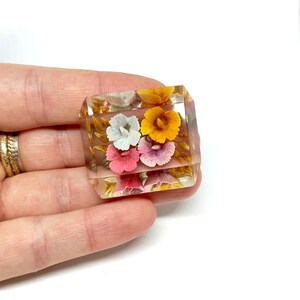 May include: A square clear glass brooch featuring a floral design. The flowers are pink, yellow, and white with gold accents.