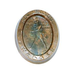 May include: An oval-shaped, bronze-colored metal object with the words "JOHN WALKER" around the edge. The center features a walking figure and the text "BORN 1840 STILL GOING STRONG".