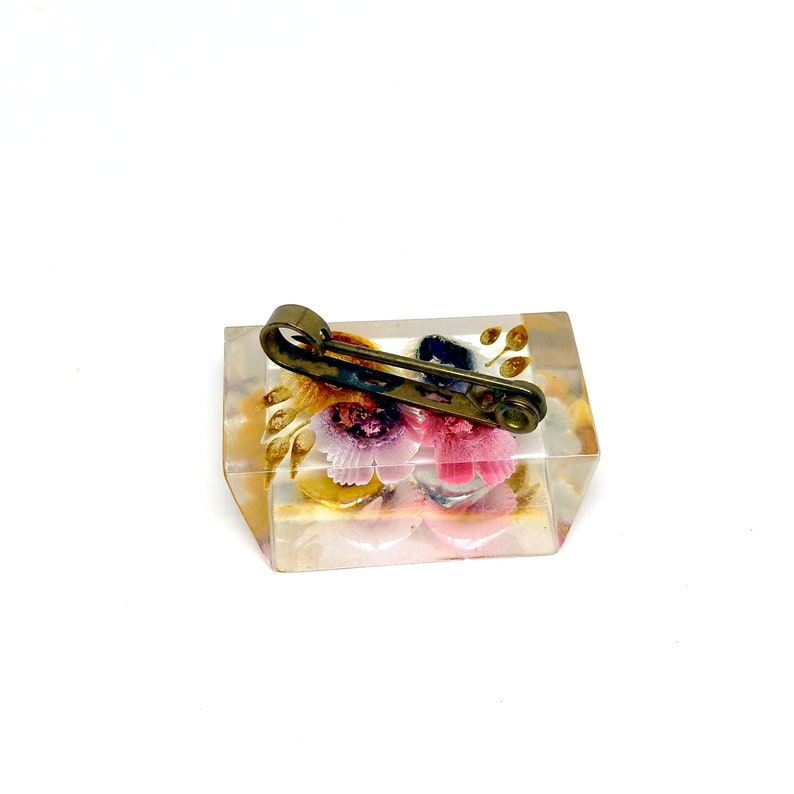 May include: A clear rectangular brooch with a floral design and a gold-toned safety pin fastening. The flowers are pink, yellow, and blue.