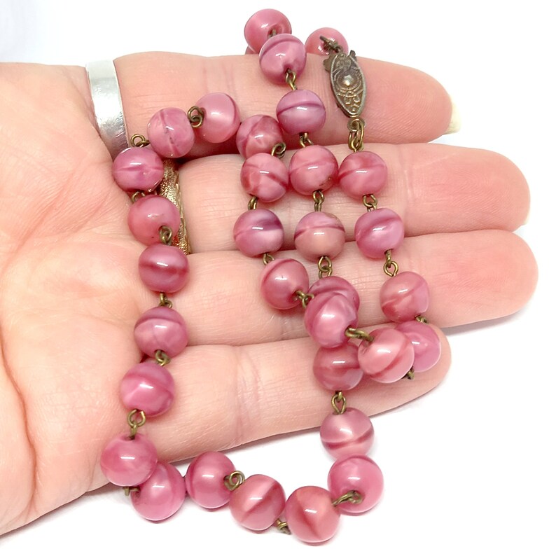 May include: A pink glass bead necklace with a metal clasp. The beads are round and have a swirl pattern.