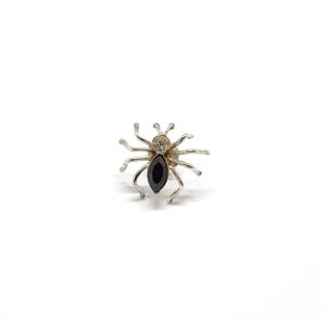 May include: A silver-toned spider brooch with a black gemstone body and clear gemstone head. The spider has eight legs and is set against a white background. This jewelry piece is a unique accessory.