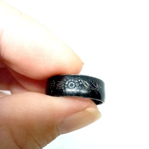 K&ouml;nnte beinhalten: Ein schwarzer Metallring mit einem silbernen Design eines Blumenmusters. Der Ring wird an einem Finger getragen.