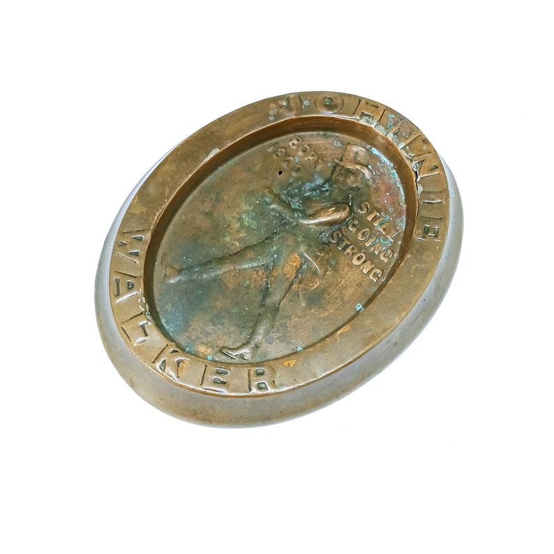 May include: An oval-shaped, bronze-colored metal object with an embossed image of a person and text. The text reads "Still Going Strong" and "Bob Heller". The object has a raised border and the word "Walker" is embossed on the bottom.