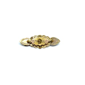 May include: A gold-toned brooch with a floral design and three pink accents. The brooch is shaped like a long oval with a heart-shaped design on each end.