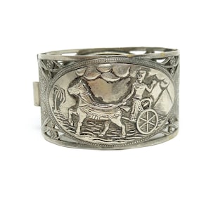 Puede incluir: Pulsera de tono plateado con un diseño ornamentado que presenta un carro tirado por un caballo y una figura con un tocado de faraón.