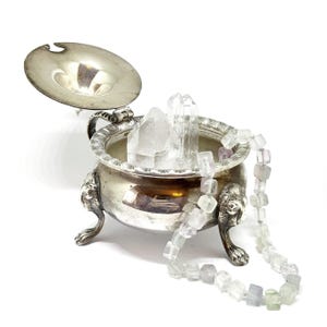 May include: A silver, footed bowl with a lid, filled with clear quartz crystals and a necklace of light green and purple beads.