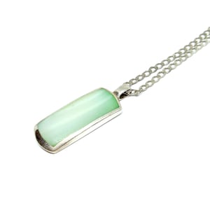 Puede incluir: Un collar de plata con un colgante rectangular. El colgante tiene una piedra translúcida verde pálido en un marco de plata, suspendido de una fina cadena de plata. El collar se muestra sobre un fondo blanco.