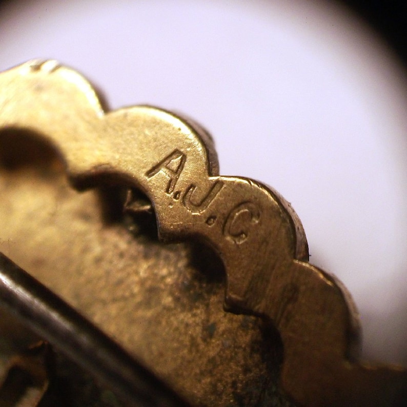 May include: Close-up of a gold-toned metal brooch with a scalloped edge. The back of the brooch is stamped with the letters "A.J.C."