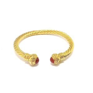 May include: A gold-coloured, open-ended bracelet with a twisted rope design. The ends feature ornate, gold-coloured caps with red, round stones. The bracelet is set against a white background.