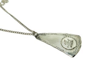 Può includere: Una collana con pendente argentato dalla forma irregolare e strutturata. Il pendente presenta un emblema circolare con un design dettagliato. La catena è un delicato stile a maglie. La collana è esposta su sfondo bianco.