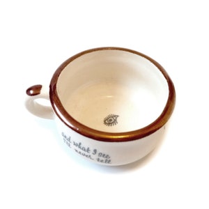 May include: White ceramic teacup with a gold rim and a black eye design inside. The cup has the text "and what I see I'll never tell" written on the side.