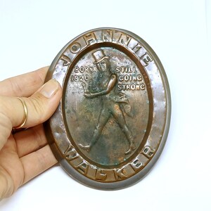 May include: Oval-shaped bronze plaque featuring the Johnnie Walker Striding Man logo. The plaque has the words "JOHNNIE WALKER" and "BORN 1820 STILL GOING STRONG" embossed around the edges. The figure is depicted in a top hat and coat.