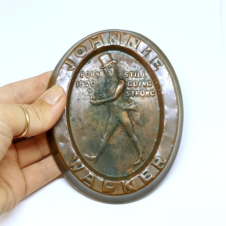 May include: Oval-shaped bronze plaque featuring the Johnnie Walker Striding Man logo. The plaque has the words "JOHNNIE WALKER" and "BORN 1820 STILL GOING STRONG" embossed around the edges. The figure is depicted in a top hat and coat.
