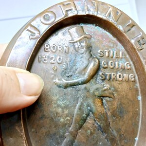 May include: A bronze-colored oval plaque with the words "JOHN WALKER" around the edge. The plaque features a relief of a man in a top hat and the text "BORN 1820" and "STILL GOING STRONG".