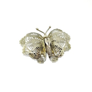 May include: A silver butterfly brooch with detailed filigree work. The wings display delicate open patterns, and the body has a textured design. This decorative brooch is designed to add a touch of elegance to clothing or accessories.