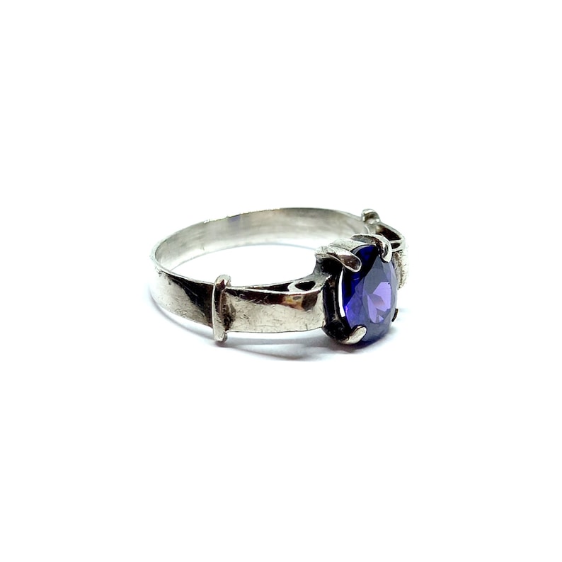 May include: A silver ring with a purple gemstone set in a prong setting. The ring has a simple band with a decorative detail on one side.