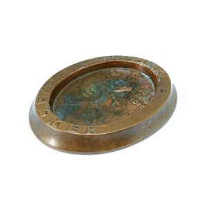 May include: Oval-shaped, antique bronze ashtray with the words "WE ARE STILL GOING STRONG" embossed around the rim. The ashtray has a shallow, concave interior with signs of age and use, and a raised, rounded edge.