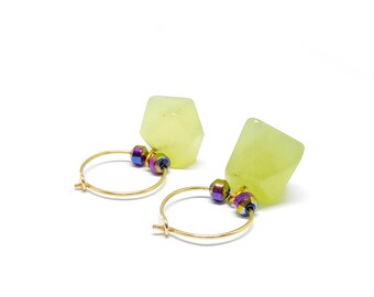 May include: A pair of gold hoop earrings with pale yellow, faceted gemstone pendants. Each earring features a small, iridescent bead and a larger, geometric gemstone. The earrings are set against a white background.