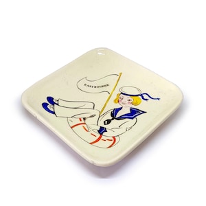 Op de afbeelding: Crèmekleurige, vierkante keramische schaal met een nautische illustratie. Het ontwerp toont een cartoonmatroos die een vlag vasthoudt met de tekst "EASTBOURNE". De matroos draagt een blauw-wit uniform en rust op een reddingsboei.