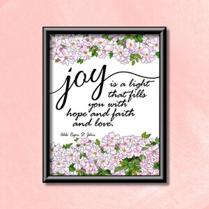 Puede incluir: Una impresión enmarcada en blanco con un borde floral. La impresión presenta una cita sobre la alegría, "La alegría es una luz que te llena de esperanza, fe y amor." La cita se atribuye a Adela Rogers St. Johns.