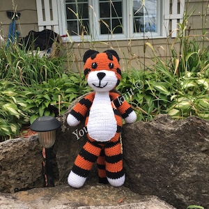 Könnte beinhalten: Ein gehäkeltes Stofftier in Form eines Tigers mit orange-schwarzen Streifen, weißen Akzenten und einem freundlichen Gesicht. Der Tiger steht auf einem Stein.