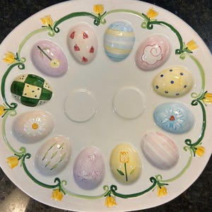 May include: A white ceramic Easter egg serving platter with a decorative border of green vines and yellow flowers. The platter features 12 egg-shaped indentations, each with a unique painted design in pastel colors.
