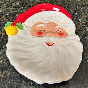 May include: A ceramic dish shaped like a smiling Santa Claus face. The figure features a bright red hat, a large white beard with textured details, rosy cheeks, and a small yellow bell with green holly leaves attached to the side of the hat.