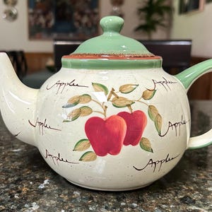 May include: A cream-colored ceramic teapot with a crackle glaze finish, featuring a hand-painted design of two red apples with green leaves. The word Apple is written in cursive script around the body. It has a light green lid and handle.