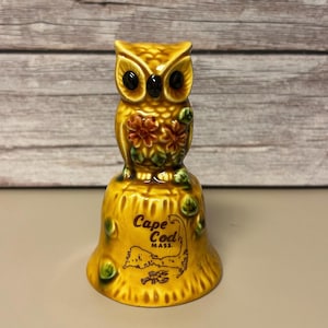 May include: A ceramic owl-shaped bell in shades of yellow and brown. The owl has large black eyes and is perched atop a bell-shaped base. The base features the text "Cape Cod MASS." and a map outline.